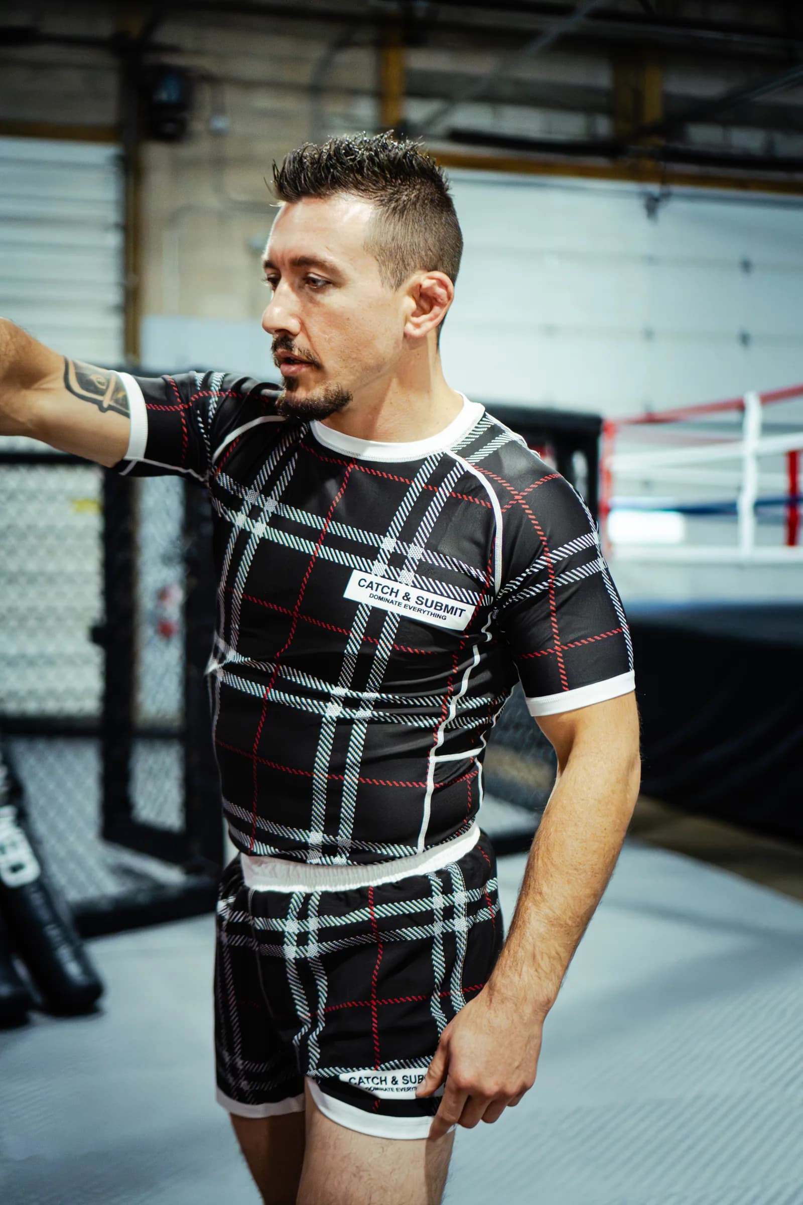 An athlete in a Catch & Submit plaid rash guard and fight shorts beside the cage